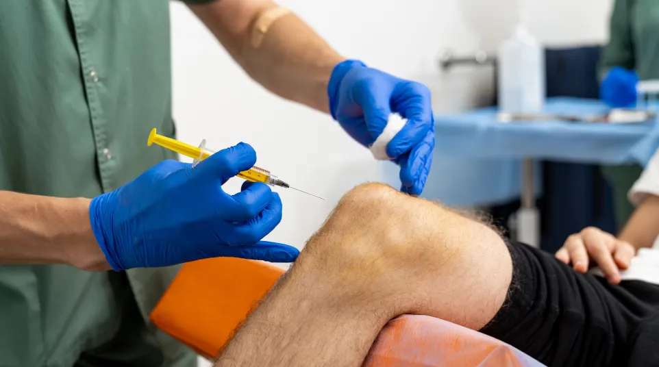 Medical professional with blue gloves administering a knee injection to a patient resting on an orange chair.
