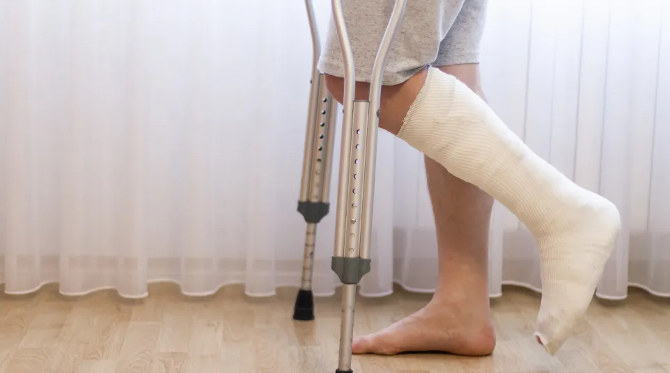 Person with a cast on foot using crutches standing on wooden floor in front of sheer curtains
