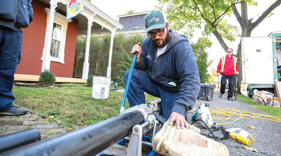Technician using pipe fusion machine to join black plastic pipes outdoors near residential area.