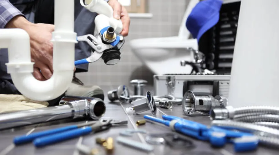 Plumber installing a white PVC pipe with various plumbing tools and fittings on a bathroom floor near a toilet.