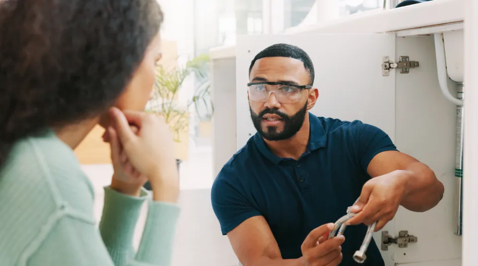 Plumber wearing safety glasses explaining a repair to a woman while fixing pipes under a sink.