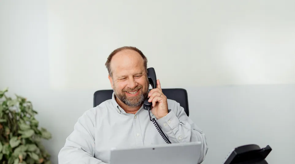 Smiling businessman talking on office phone while working on laptop at desk with notebook and coffee cup