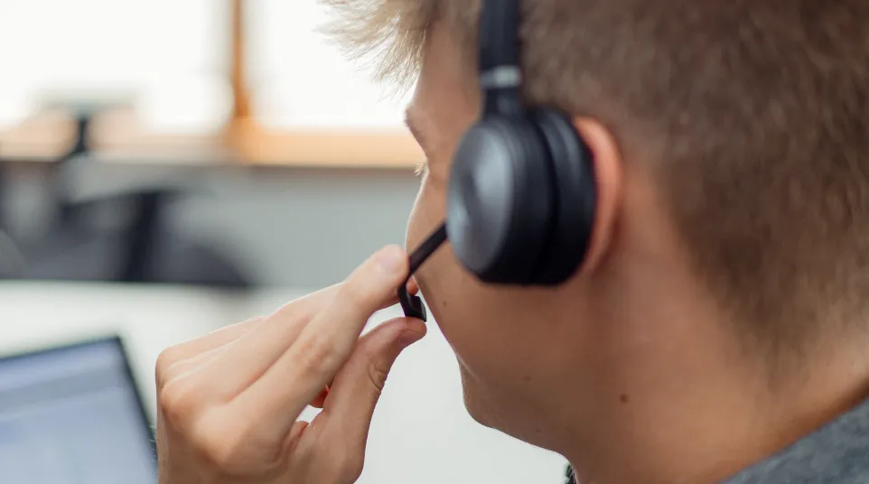 Young man wearing headset with microphone working on laptop in bright office environment.