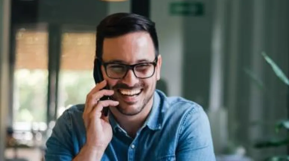 Smiling man in glasses talking on phone at desk with laptop and notebook in modern office.