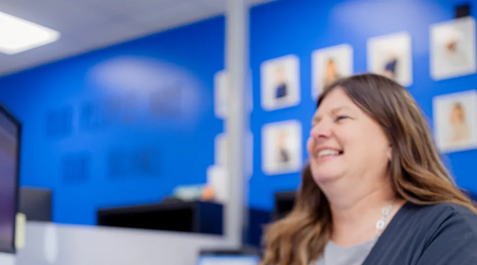 Woman smiling while typing on a keyboard at a white desk in a blue office with framed photos on the wall.