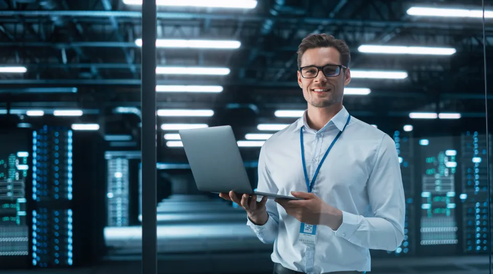 Smiling man in a modern data center holds a laptop and poses confidently for the camera.