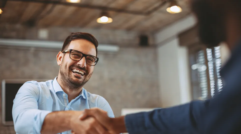 Two professionals shaking hands in a modern office, smiling and engaged in a positive interaction.