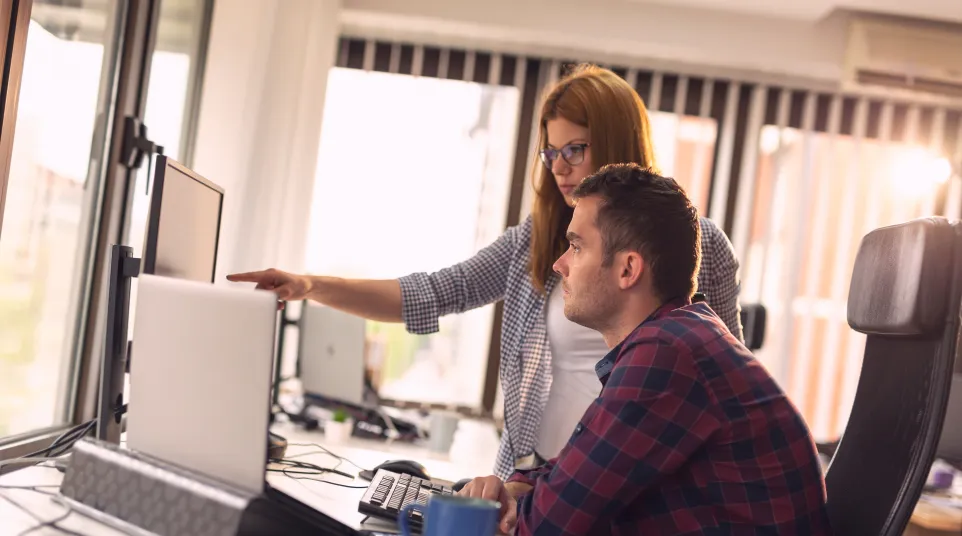 Two colleagues collaborating at a desk with computers in a bright office setting.