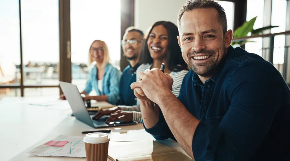A diverse team collaborates at a table with laptops, notes, and coffee, smiling and engaged.