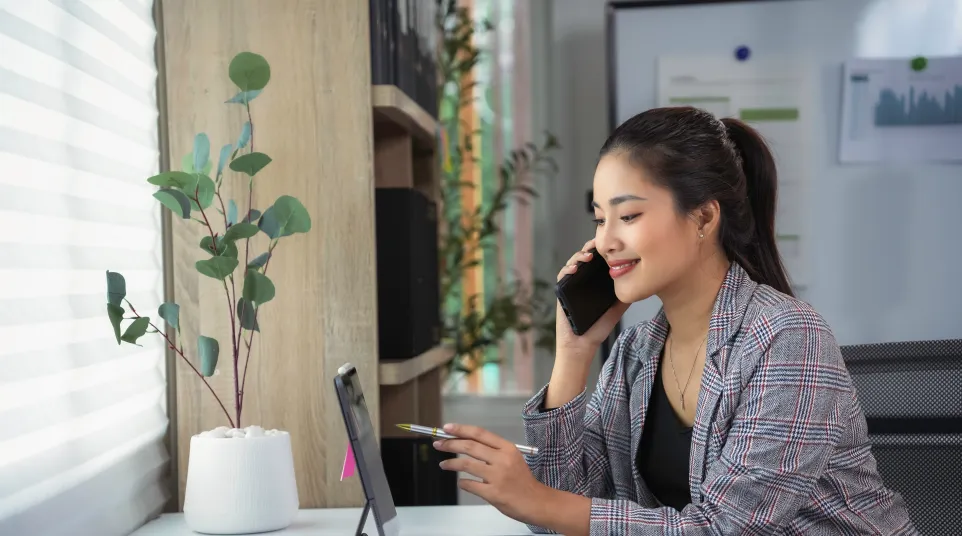Woman talking on phone while working on tablet in modern office space with plants and bright natural light.