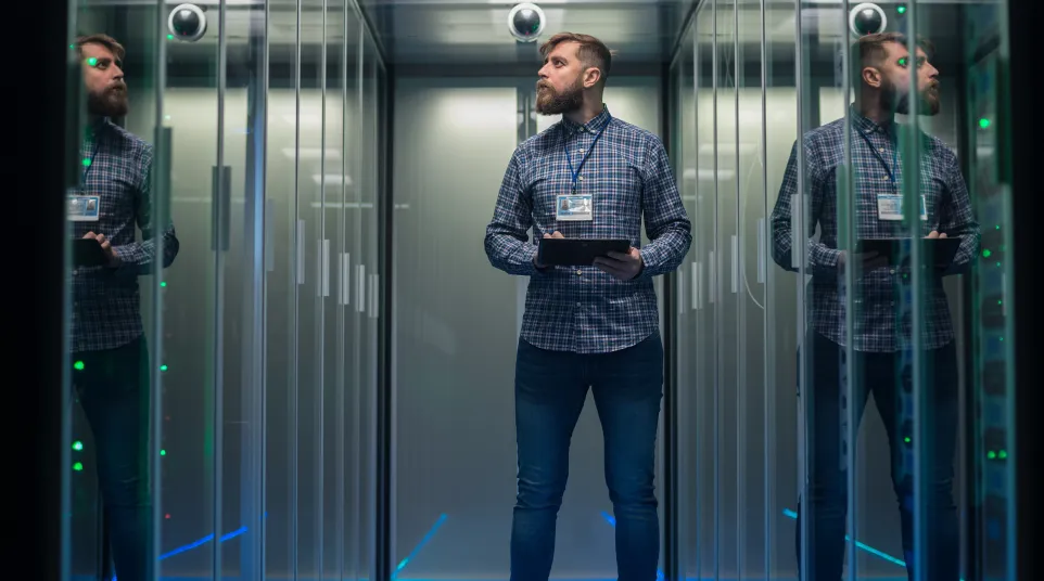 A tech professional holds a tablet in a server room surrounded by reflective glass panels.