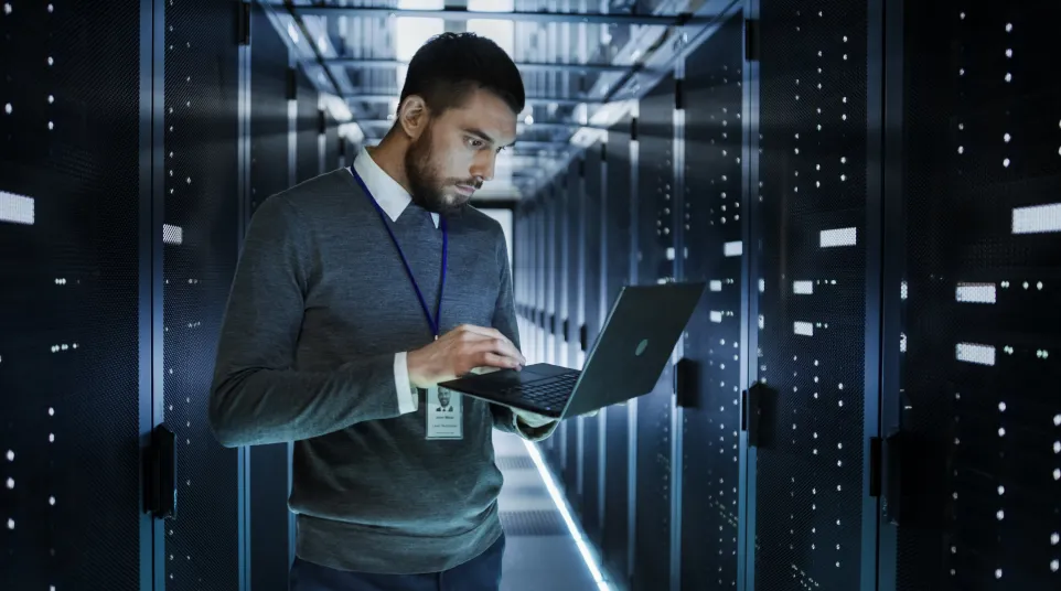 A focused man using a laptop in a data center surrounded by server racks.