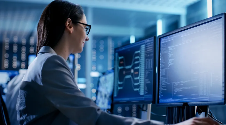 Professional woman working on dual monitors with coding data in a modern tech environment.