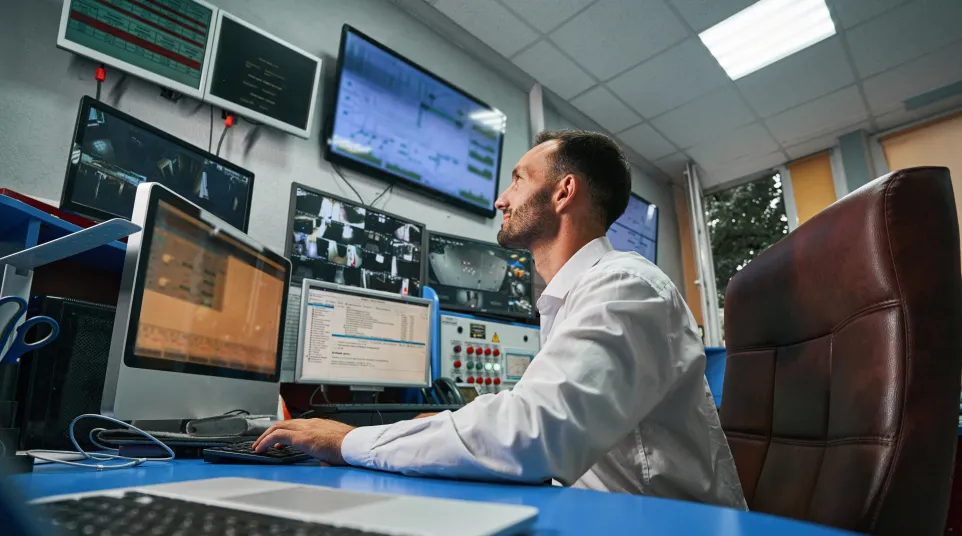 Man working at a computer in a modern control room with multiple screens displaying data.