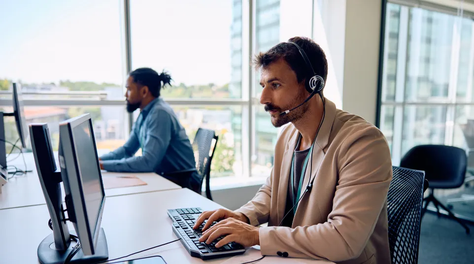 A man with a headset working at a desk with computers in a modern office environment.