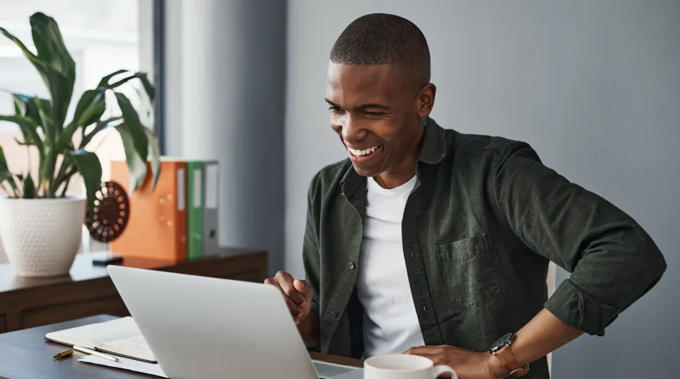 A man smiling while working on a laptop at a desk, with plants and a cup nearby.