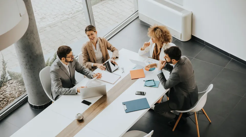 Four professionals in a modern meeting room discussing plans over a table with laptops and documents.