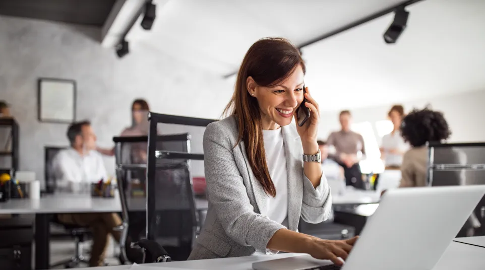 Woman smiling while talking on phone and working on laptop in a modern office setting.