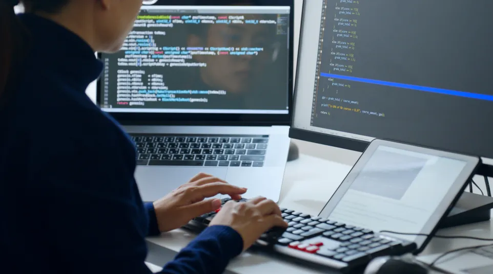 A woman coding on a laptop and monitor with visible lines of code, creating software solutions.
