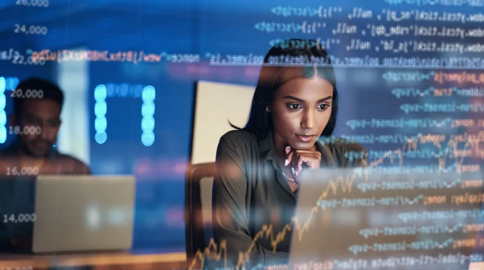 Focused woman analyzing data on a laptop with coding graphics overlay in a modern office.