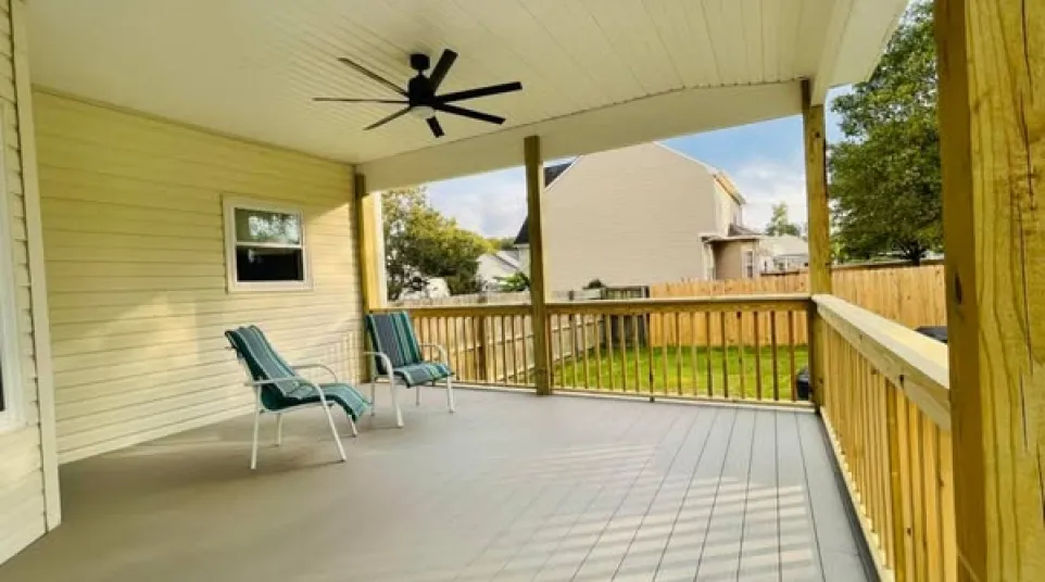 Spacious covered porch with ceiling fan, two green chairs, and wooden railing overlooking backyard.
