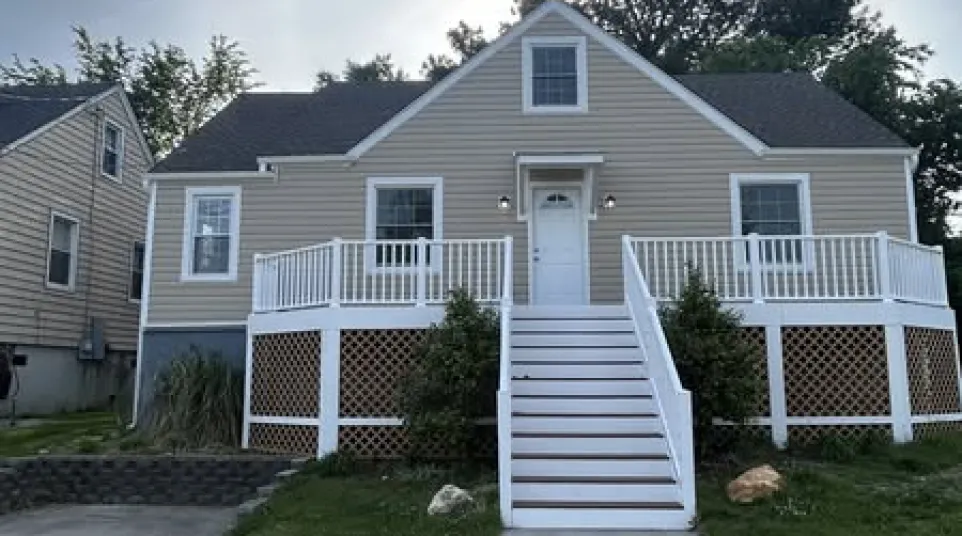 Beige raised house with white railings and stairs surrounded by green lawn under a cloudy sky.