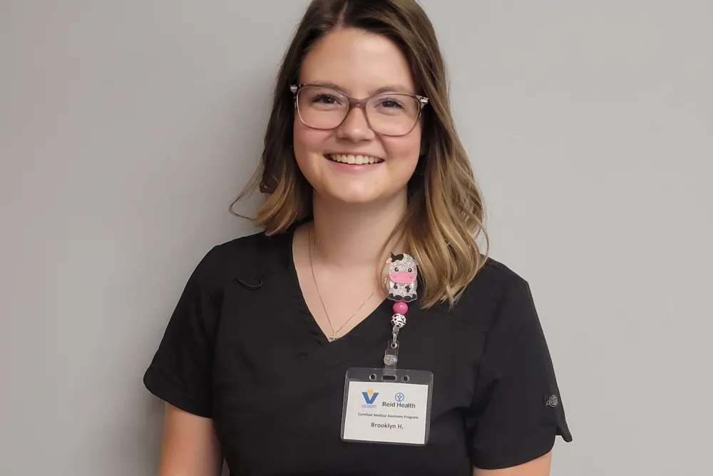 Smiling Reid Health CMA program participant wearing black scrubs and glasses with a Reid Health name badge on gray background