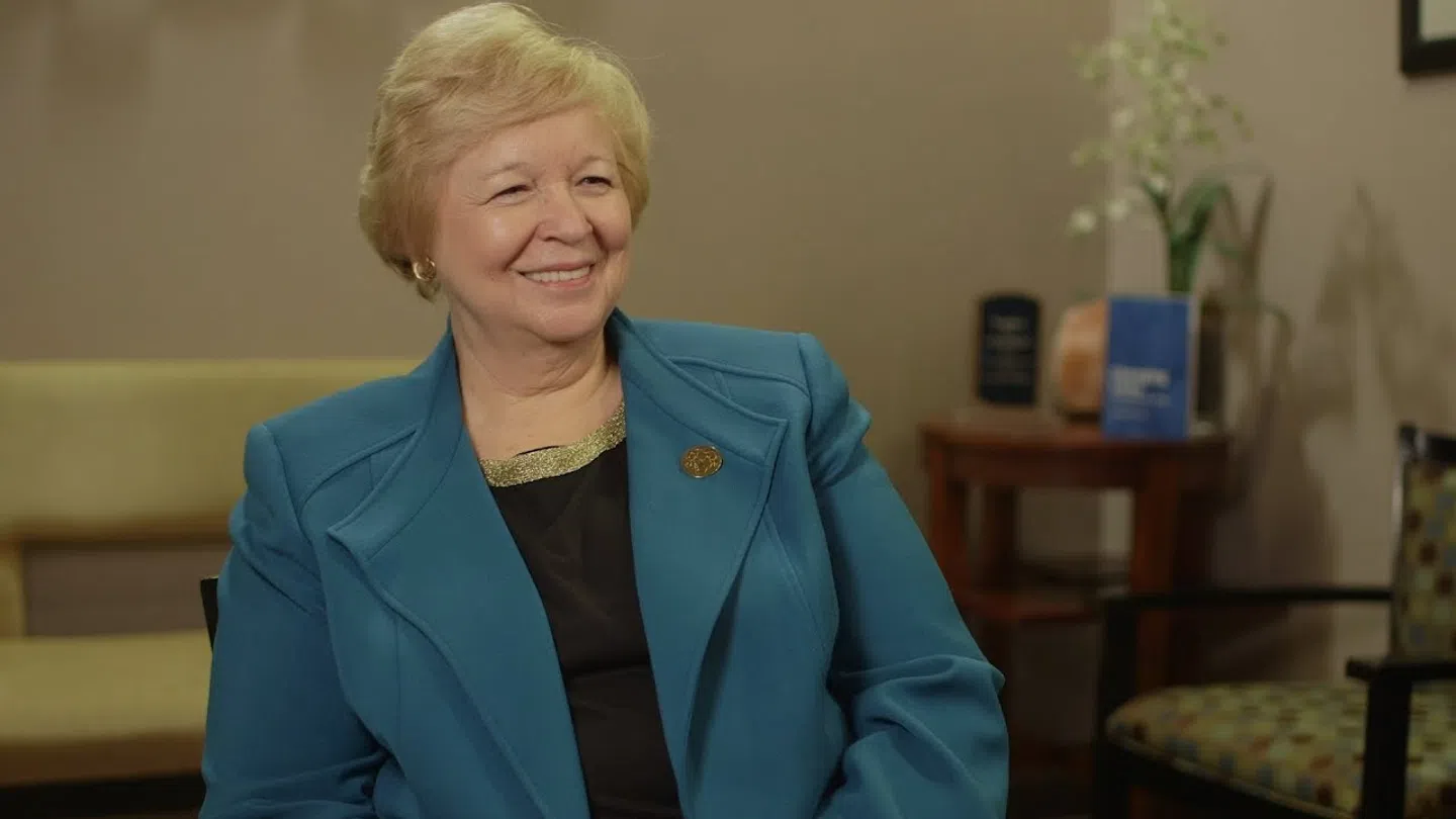 Smiling older woman in blue blazer sitting in a warmly lit room with chairs and a small table in the background
