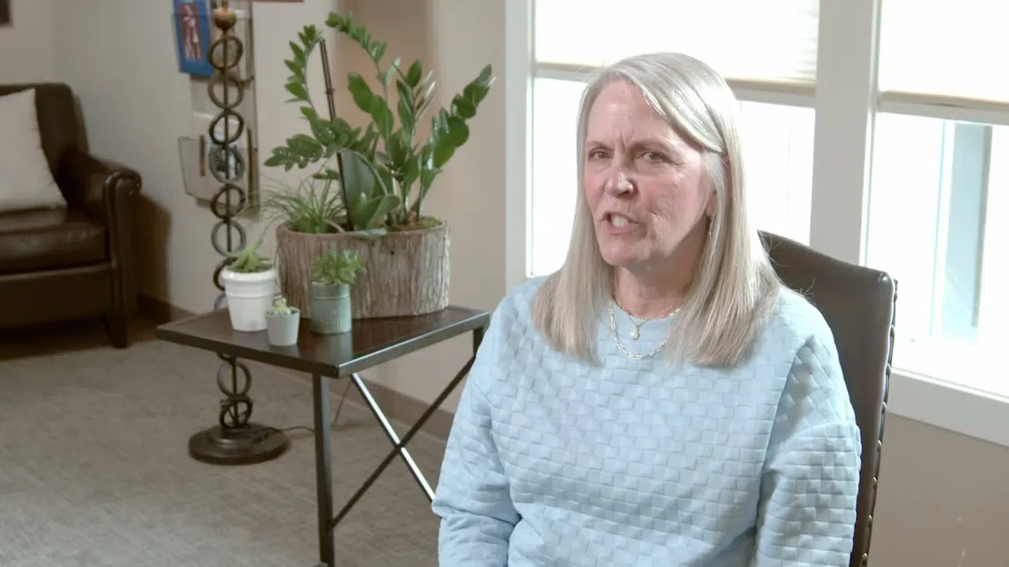Middle-aged woman with long gray hair sitting in a chair in a bright room with plants and a sofa.