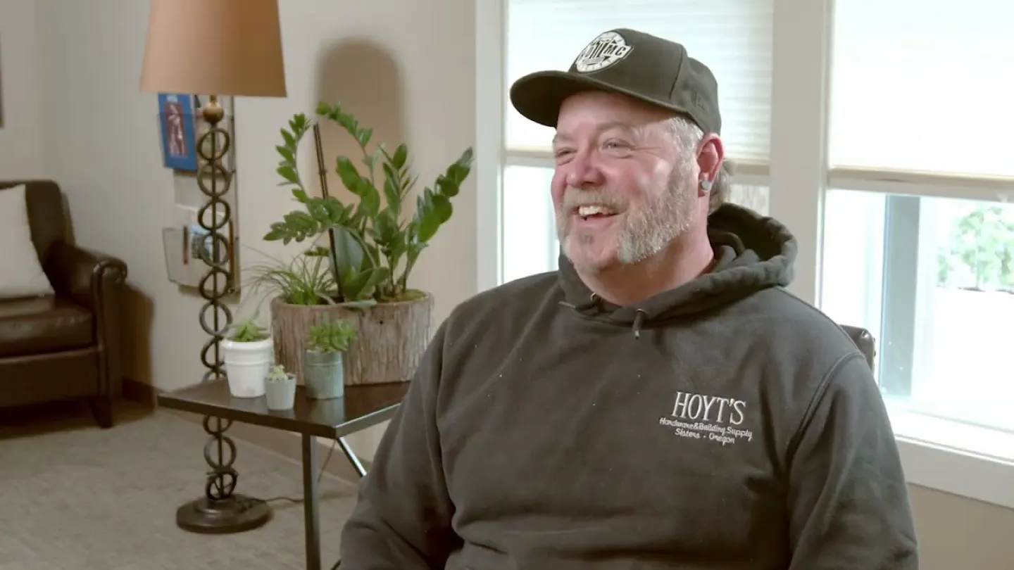 Smiling man in casual hoodie and cap sitting indoors near plants and window with natural light.