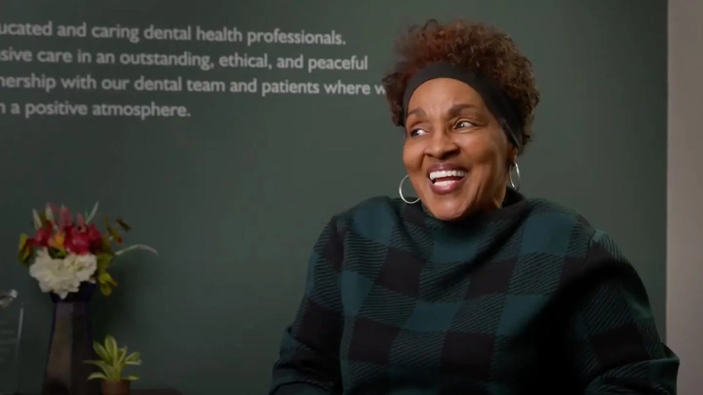 Smiling woman in checkered sweater sitting in a room with inspiring dental care text and flower vase.
