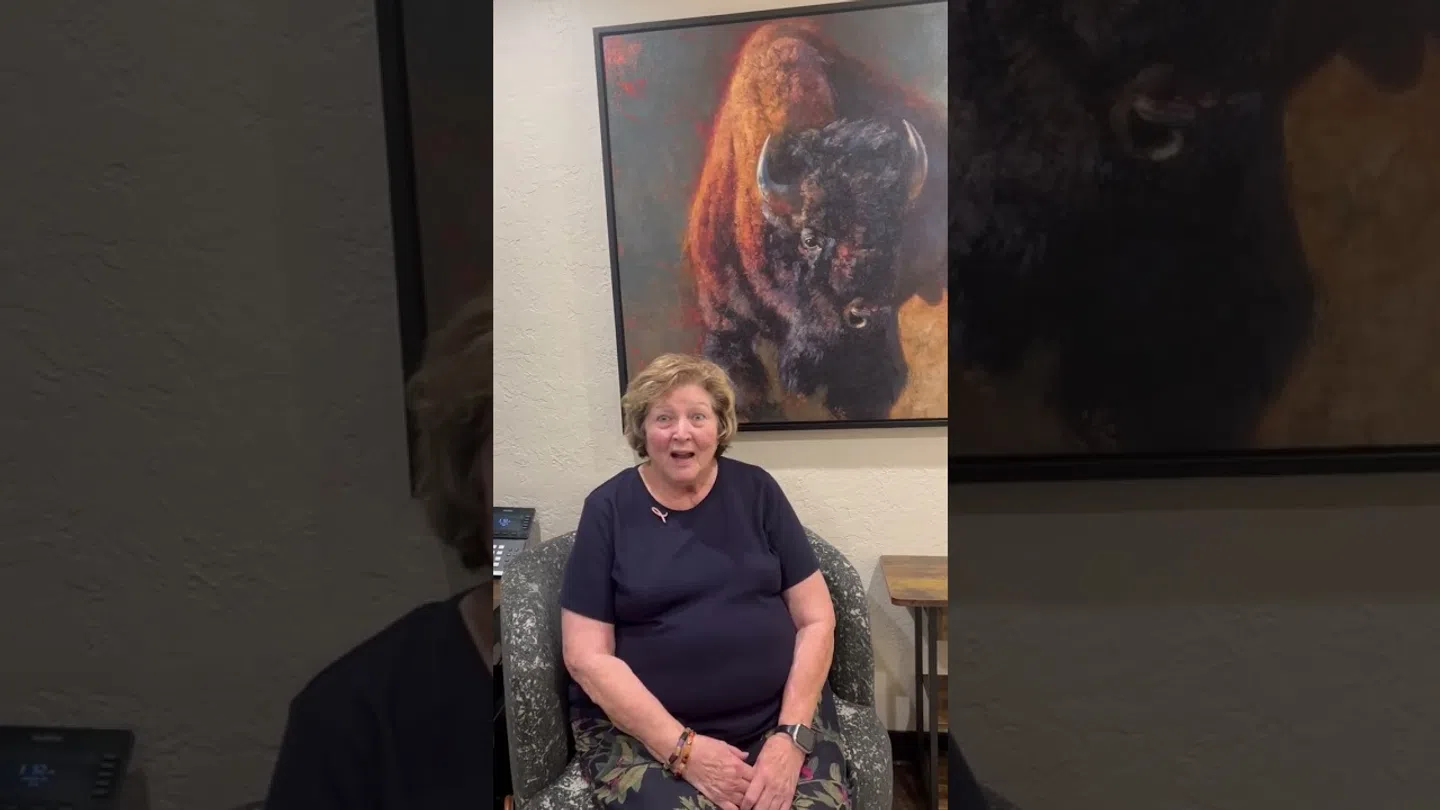 Smiling woman seated in armchair under large painting of a bison with warm tones on wall.