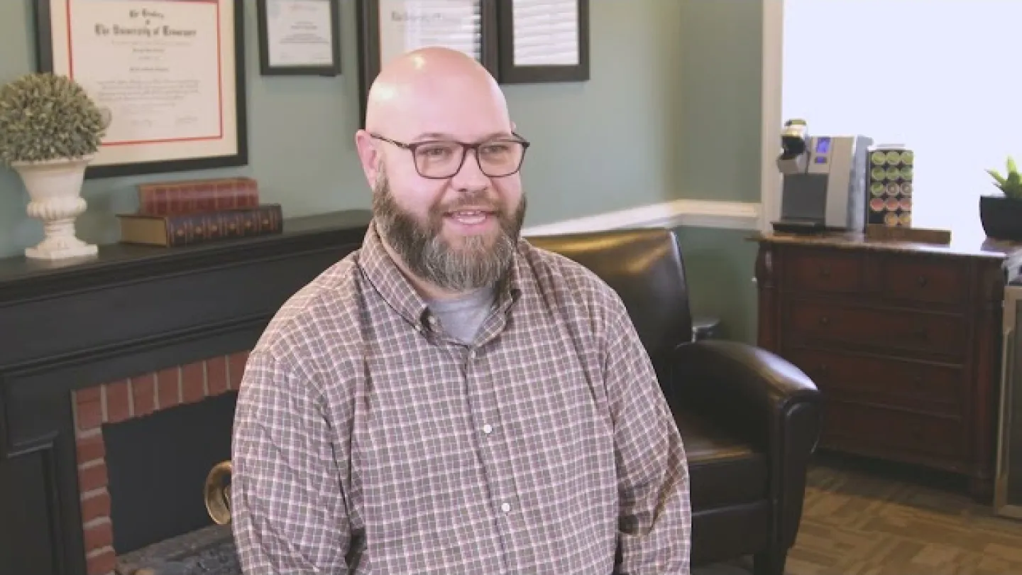 Smiling bearded man with glasses in plaid shirt sitting in cozy office with certificates and bookshelf.