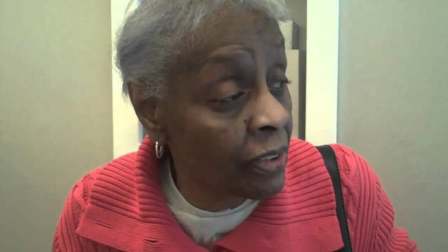 Elderly woman with short gray hair wearing a red sweater and looking to the side in an indoor setting