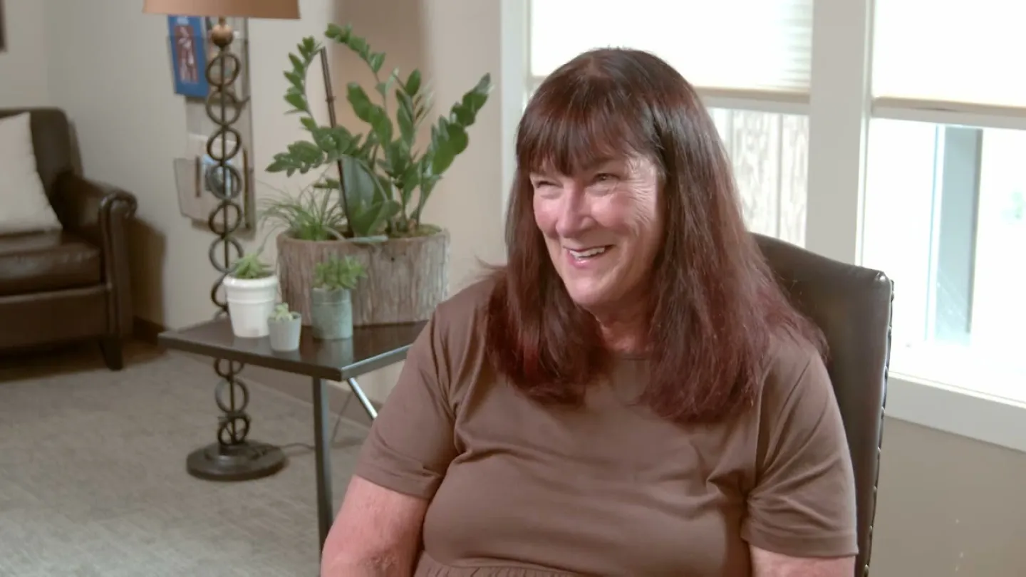 Smiling woman with brown hair seated in a cozy living room with plants and natural light.