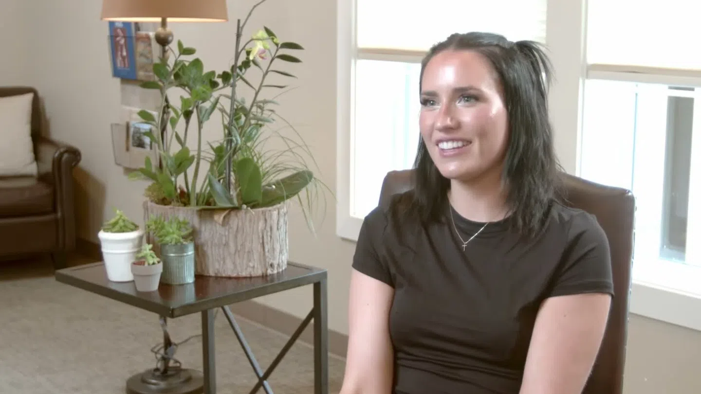 Smiling young woman with dark hair sitting in a chair indoors near a table with potted plants and natural light.