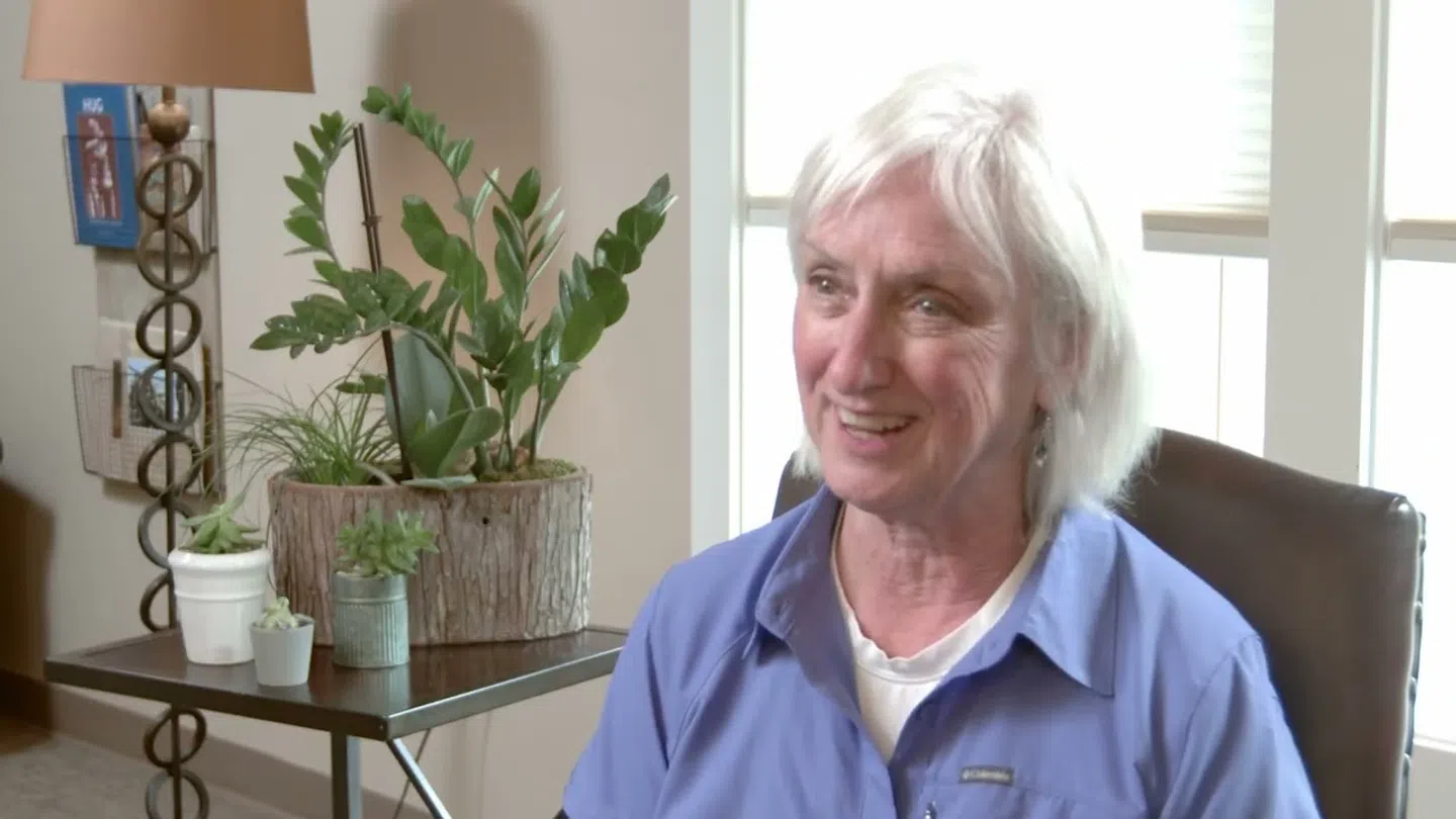 Smiling older woman with white hair sitting indoors near window and green potted plants on side table.