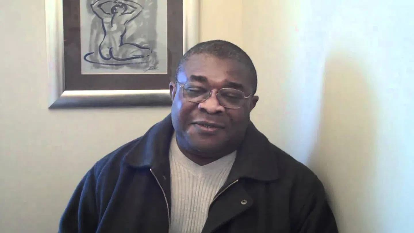 Man wearing glasses and a dark jacket sitting indoors with framed abstract art on the wall behind him