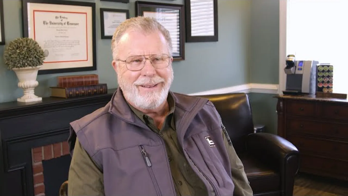 Smiling elderly man with glasses and beard sitting in office with framed diplomas and cozy decor.
