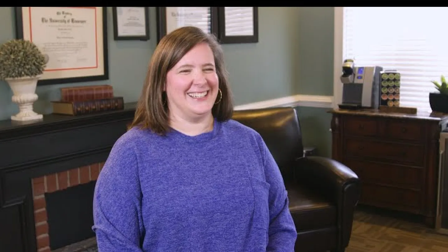 Woman smiling wearing a purple sweater sitting in a cozy office with framed certificates and dark leather chair.