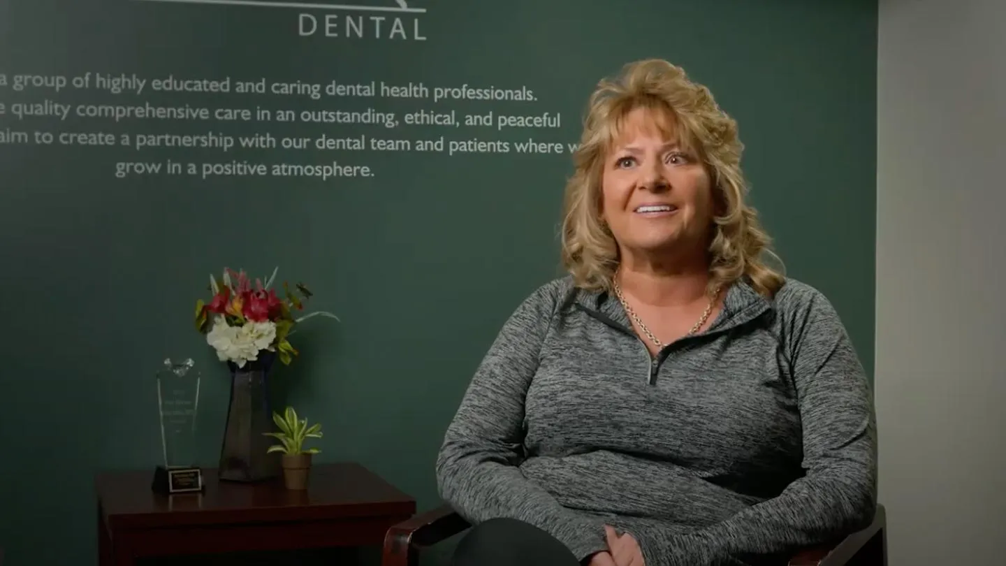 Smiling woman in gray sweater sitting in dental office with flowers and plaque on table behind her.