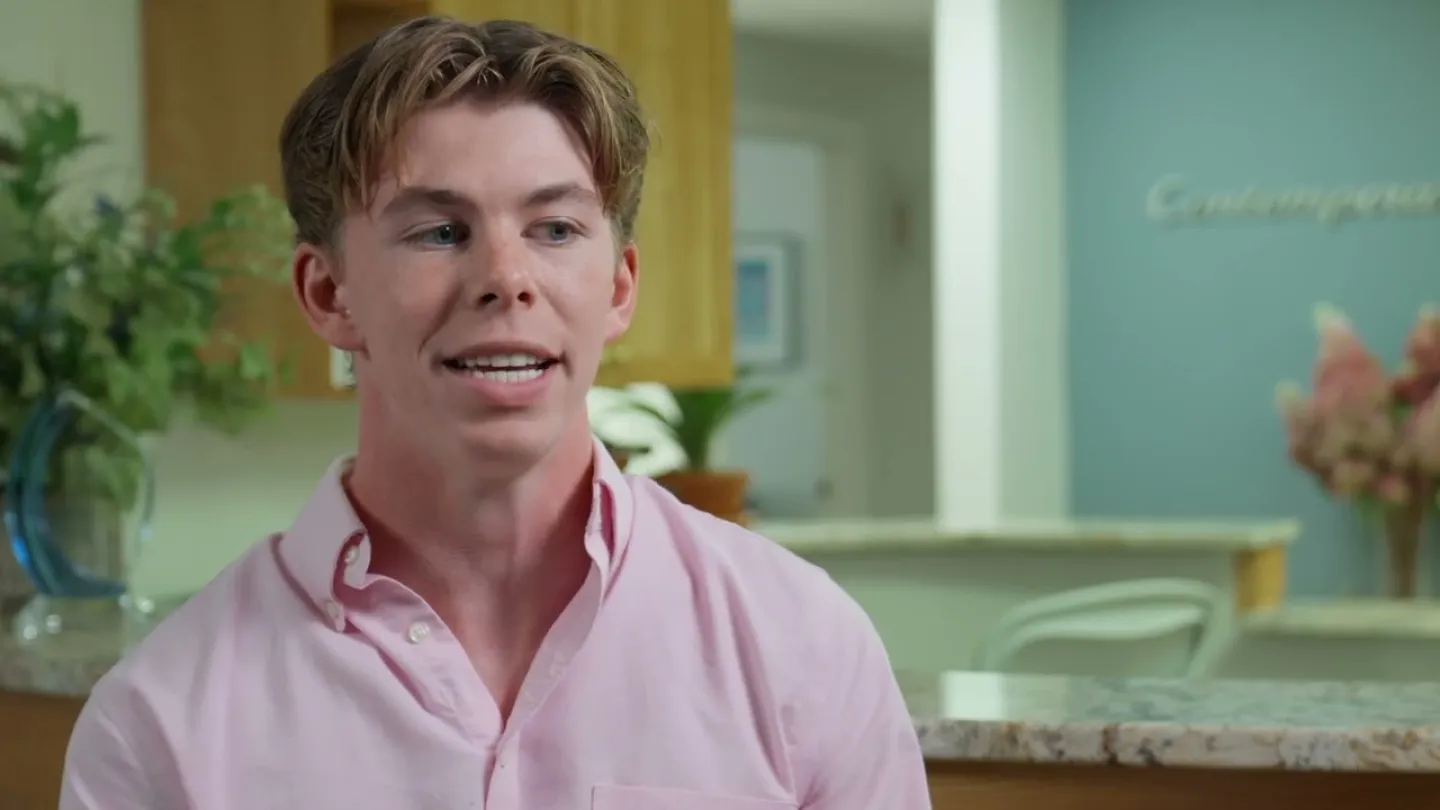 Young man in a pink shirt speaking indoors with a kitchen background and plants visible.