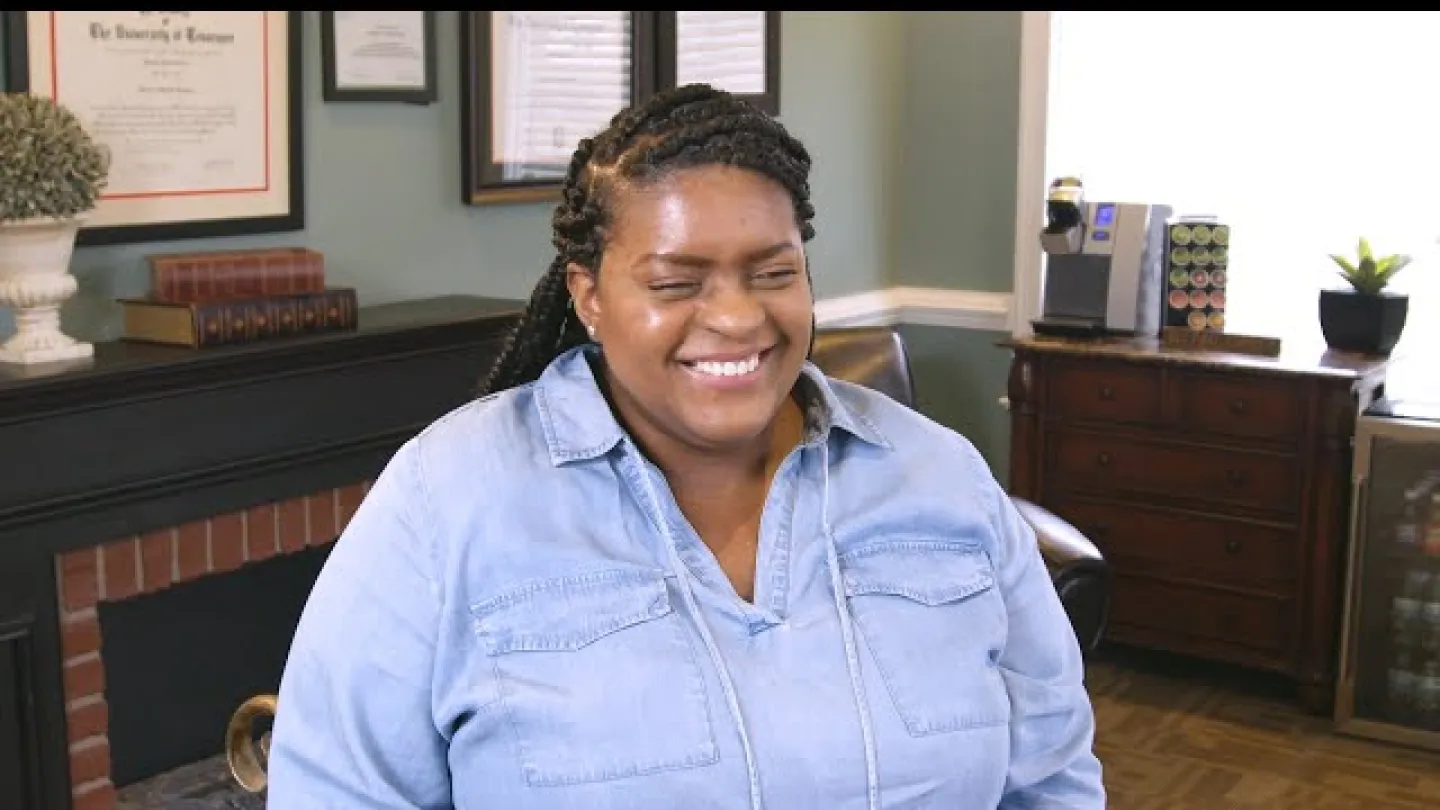Smiling woman with braided hair wearing a blue shirt sitting in a cozy office with framed certificates and wooden furniture.