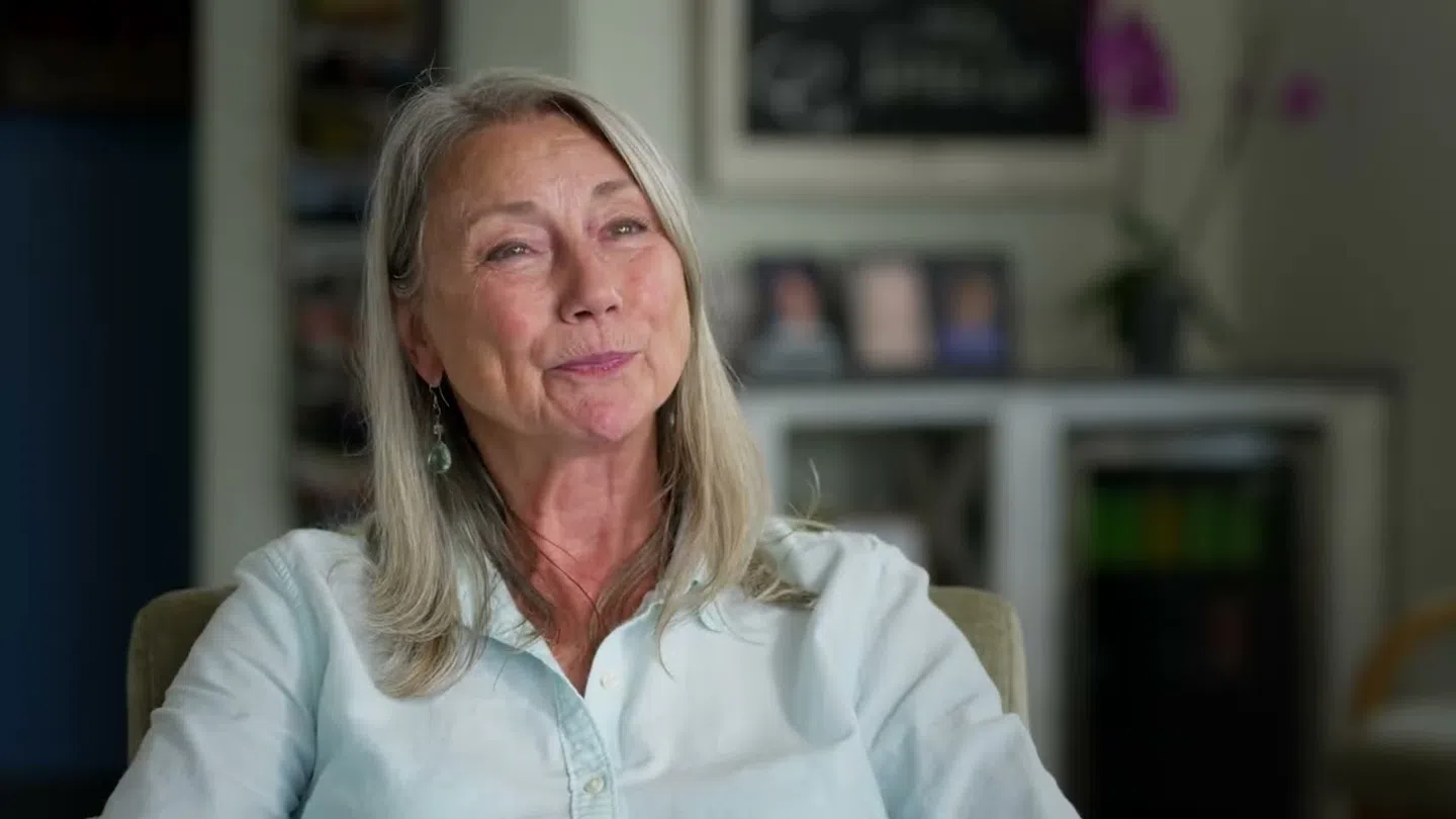 Smiling mature woman with long gray hair wearing a light blue shirt sitting in a cozy home setting.