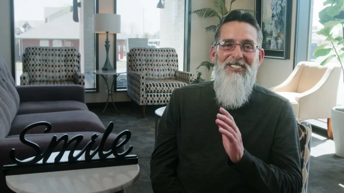 Smiling elderly man with gray beard sitting in a cozy room with modern chairs and a table sign saying Smile
