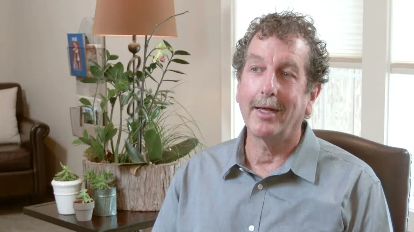 Middle-aged man with curly hair and mustache sitting in a chair in a living room with plants and a lamp.