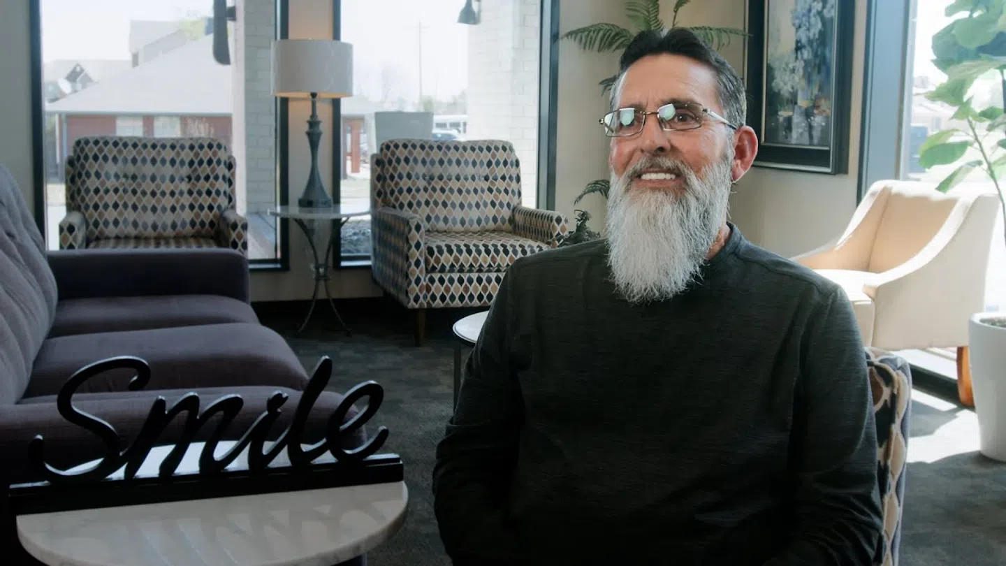Smiling elderly man with glasses and long gray beard sitting in a modern living room with natural light.