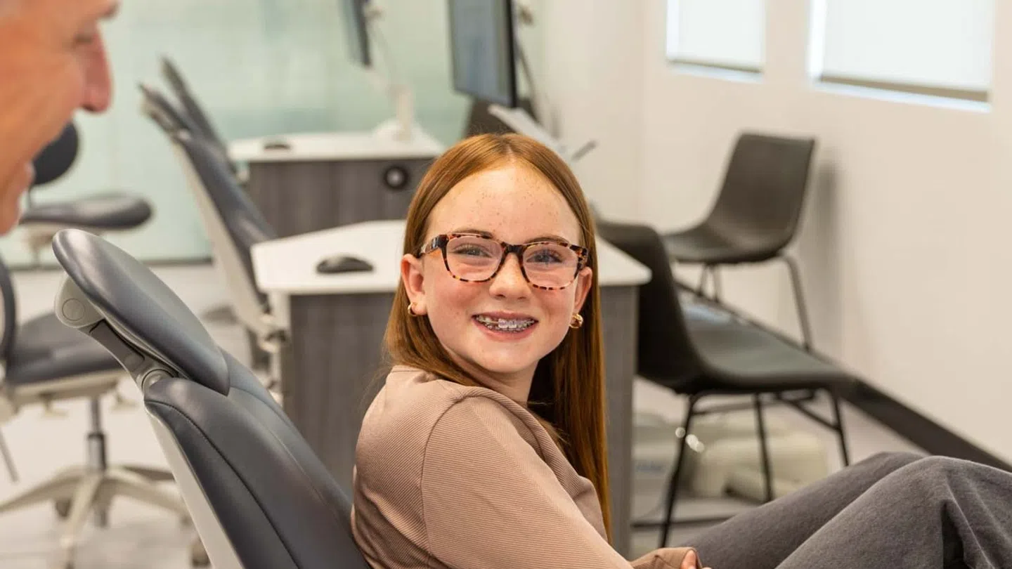 Smiling girl with braces sitting in dental chair talking to dentist in bright orthodontic office.