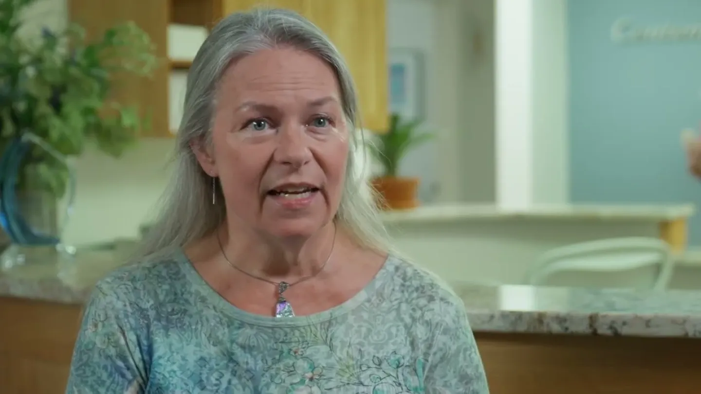 Middle-aged woman with long gray hair speaking indoors in a well-lit room with plants and kitchen background.
