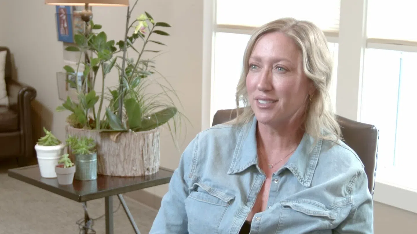 Blonde woman in a denim shirt sitting in a chair with a table of potted plants in a bright room.